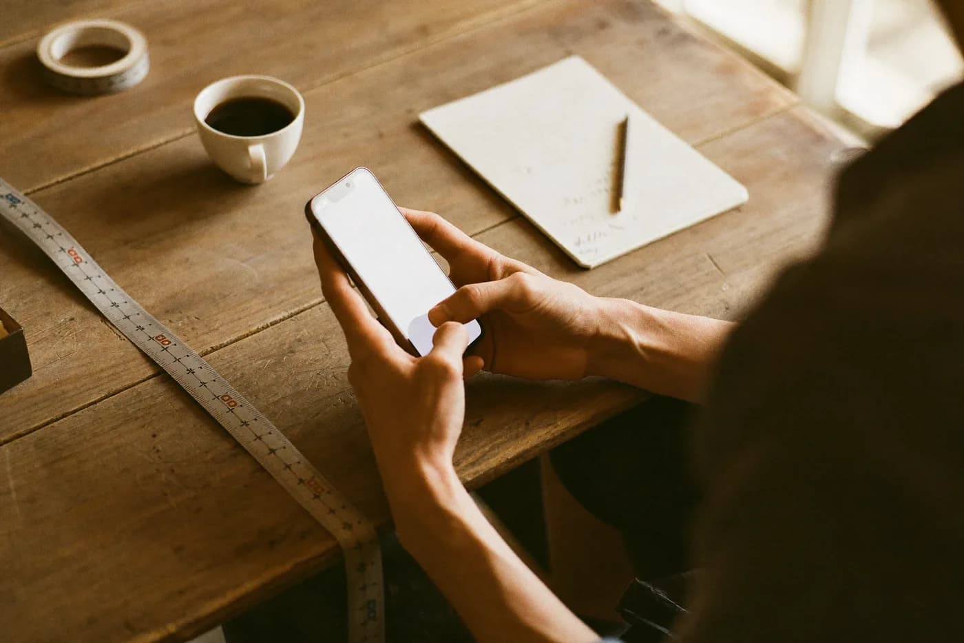 Manos sosteniendo un teléfono sobre una mesa de trabajo con una libreta y un café
