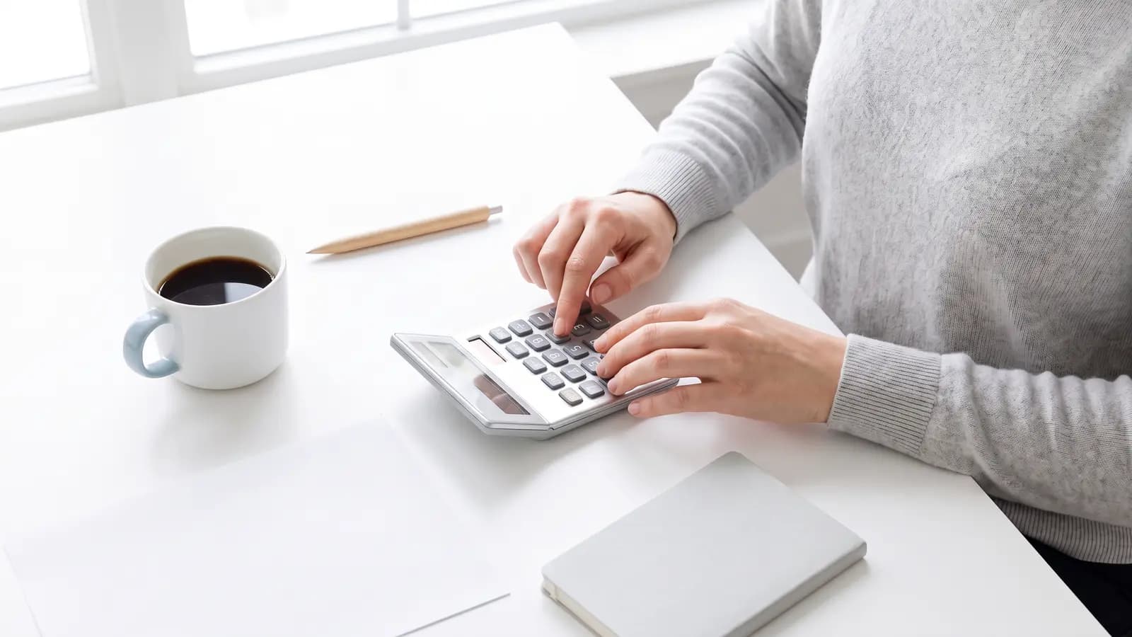 Las manos de una persona usan una calculadora sobre un escritorio blanco, junto a una hoja impresa y una taza de café.
